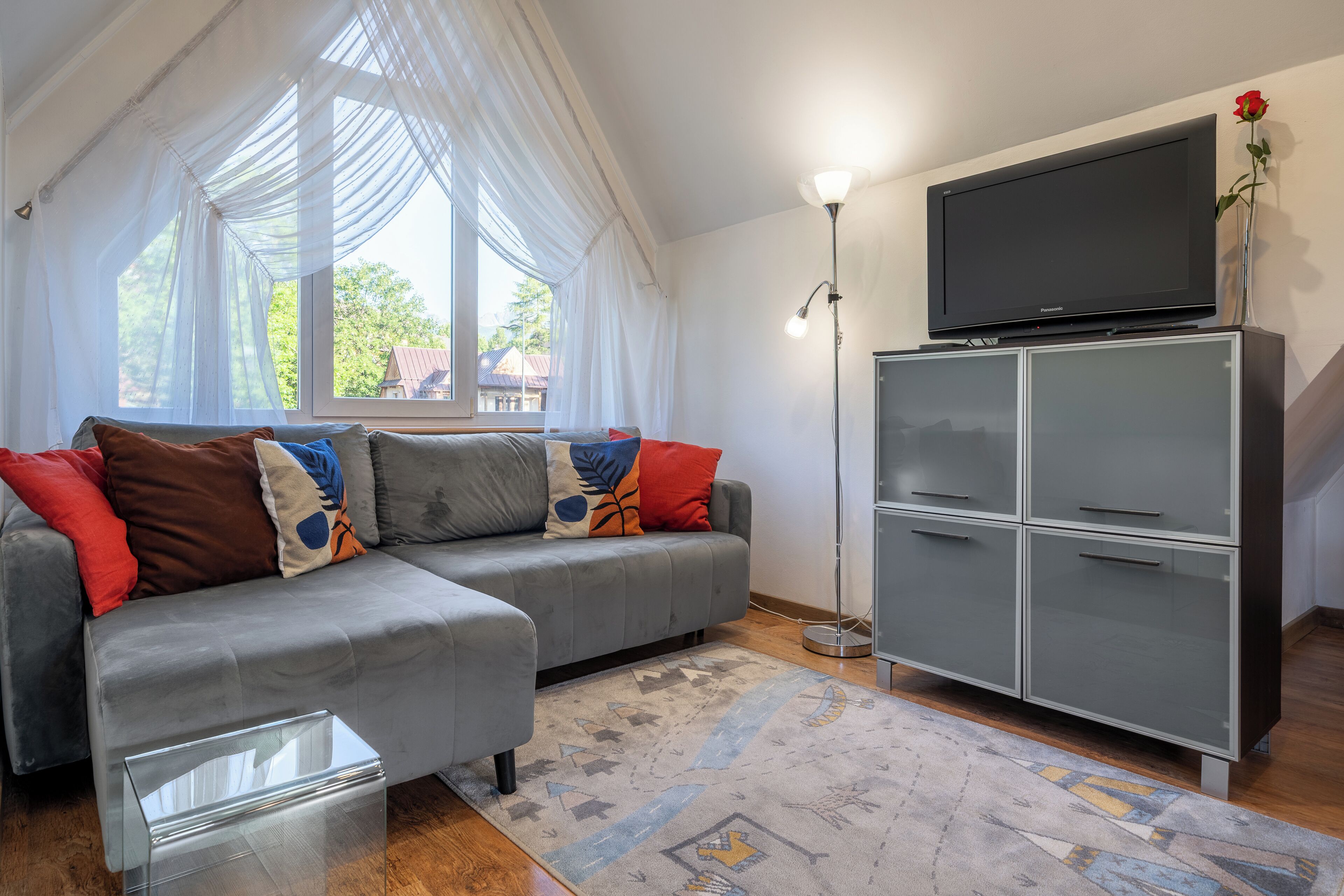 A modern living room with a grey sectional sofa, a glass coffee table, a grey cabinet with a flat-screen TV, and a patterned rug.