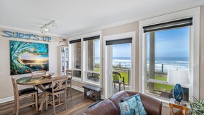 A living room with a view of the ocean, a dining area with a glass table, and a large painting with the text "SURF'S UP".