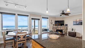 A spacious living room with a large window overlooking the ocean, a stone fireplace, and a dining area with wooden chairs and a table.