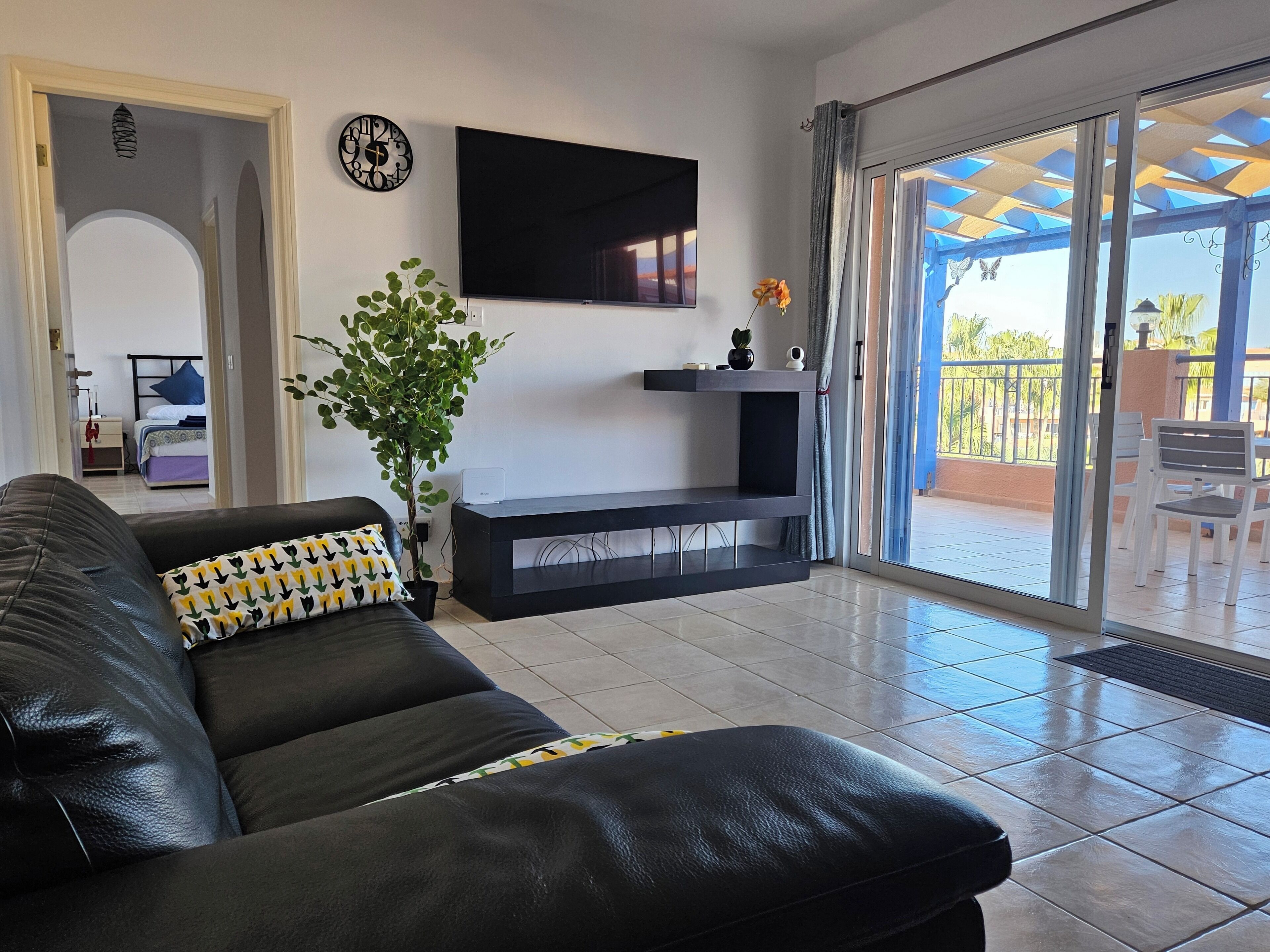 A living room with a black leather sofa, a flat-screen TV mounted on the wall, a potted plant, and a sliding glass door leading to an outdoor area with a patio and chairs.