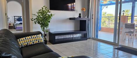 A living room with a black leather sofa, a flat-screen TV mounted on the wall, a potted plant, and a sliding glass door leading to an outdoor area with a patio and chairs.
