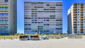Un immeuble de grande hauteur doté de nombreux balcons, situé sur une plage de sable, avec une clôture en bois en premier plan.
