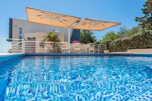 Una piscina con sillones de descanso y sombrillas, rodeada por un edificio moderno y un cielo azul despejado.