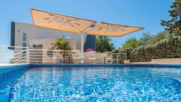 A swimming pool with lounge chairs and umbrellas, surrounded by a modern building with a clear blue sky. - Sargas Suite 5 with pool view (Novalja)