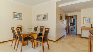 A dining area with a glass table and wooden chairs, a kitchen in the background, and a hallway leading to another room. - Ivana Apartment by HR Madeira (Funchal)