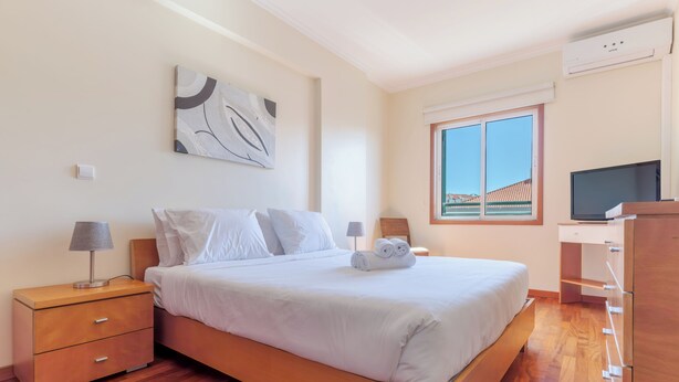 A neatly arranged bedroom with a bed, bedside tables, a lamp, a TV, and a window with a view. - Ivana Apartment by HR Madeira (Funchal)