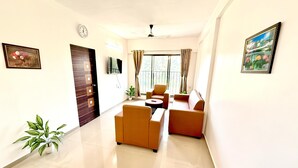 A modern living room with a sofa set, a TV mounted on the wall, and a large window with curtains. - Konkan Pearl Suites (Malvan)