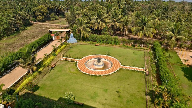 An aerial view of a landscaped area with a central circular garden, palm trees, and a statue. - Konkan Pearl Suites (Malvan)