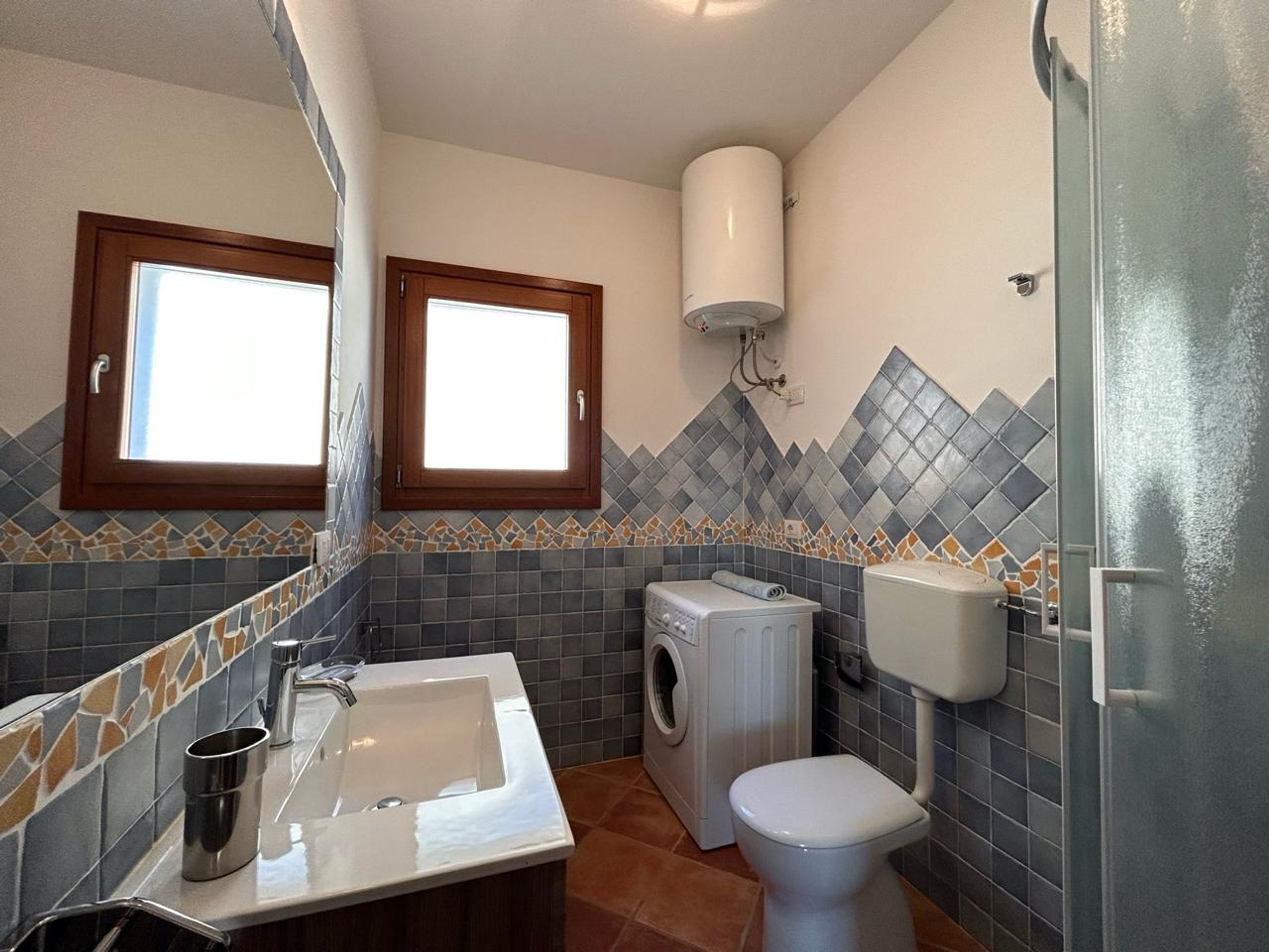 Un baño pequeño con lavamanos, inodoro y lavadora. Dos ventanas permiten la entrada de luz natural.