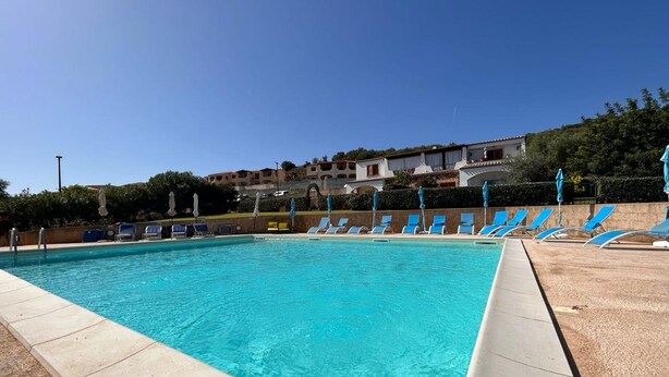 Una piscina con agua azul cristalina, rodeada de sillones de descanso y sombrillas, con edificios al fondo.