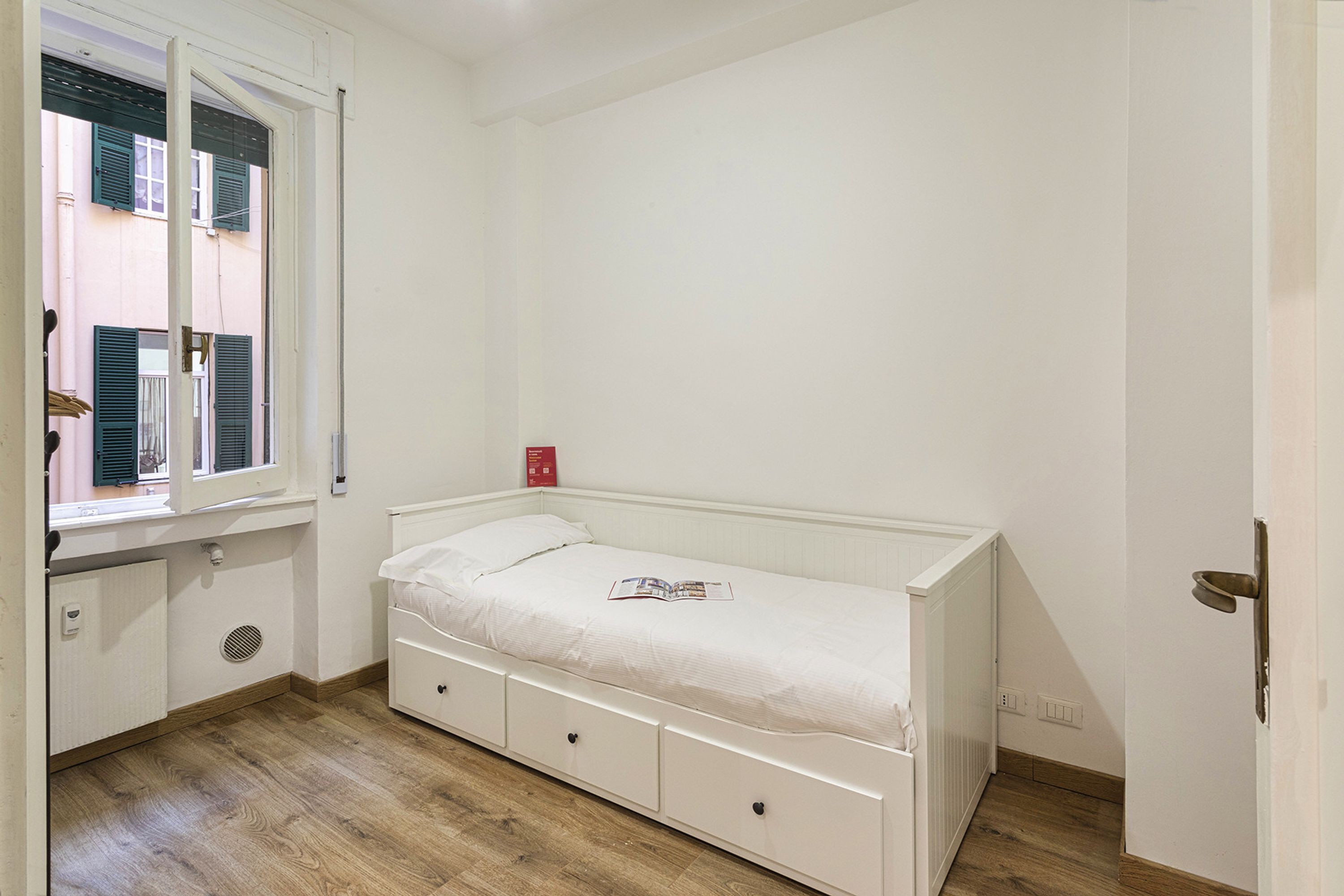 A small, white bedroom with a single bed, a window with shutters, a radiator, and a door.