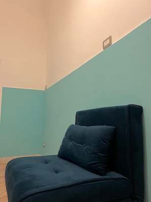 A room with a single dark blue sofa against a teal wall, a light switch, and a power outlet on the wall.