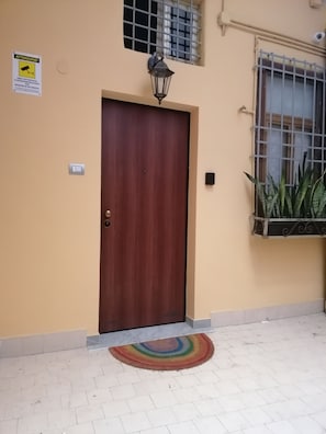 A closed wooden door with a light fixture above it, a window with a metal grill, and a potted plant on the right.