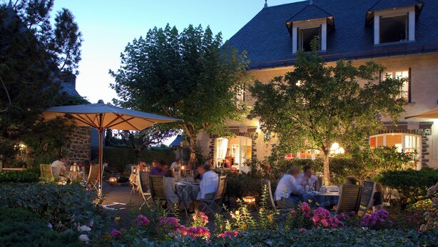 An outdoor dining area with tables and chairs, surrounded by lush greenery and flowering plants. - Le Baillage (Salers)