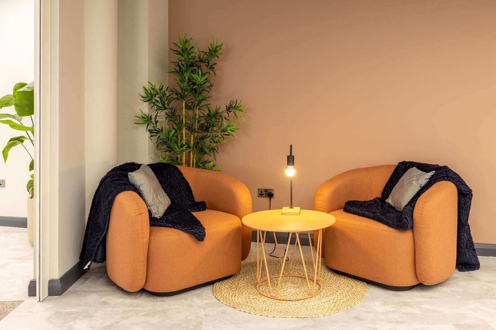 Deux fauteuils orange ornés de plaids, une petite table ronde avec une lampe et une plante en bambou dans une pièce moderne aux murs beiges et au sol carrelé.