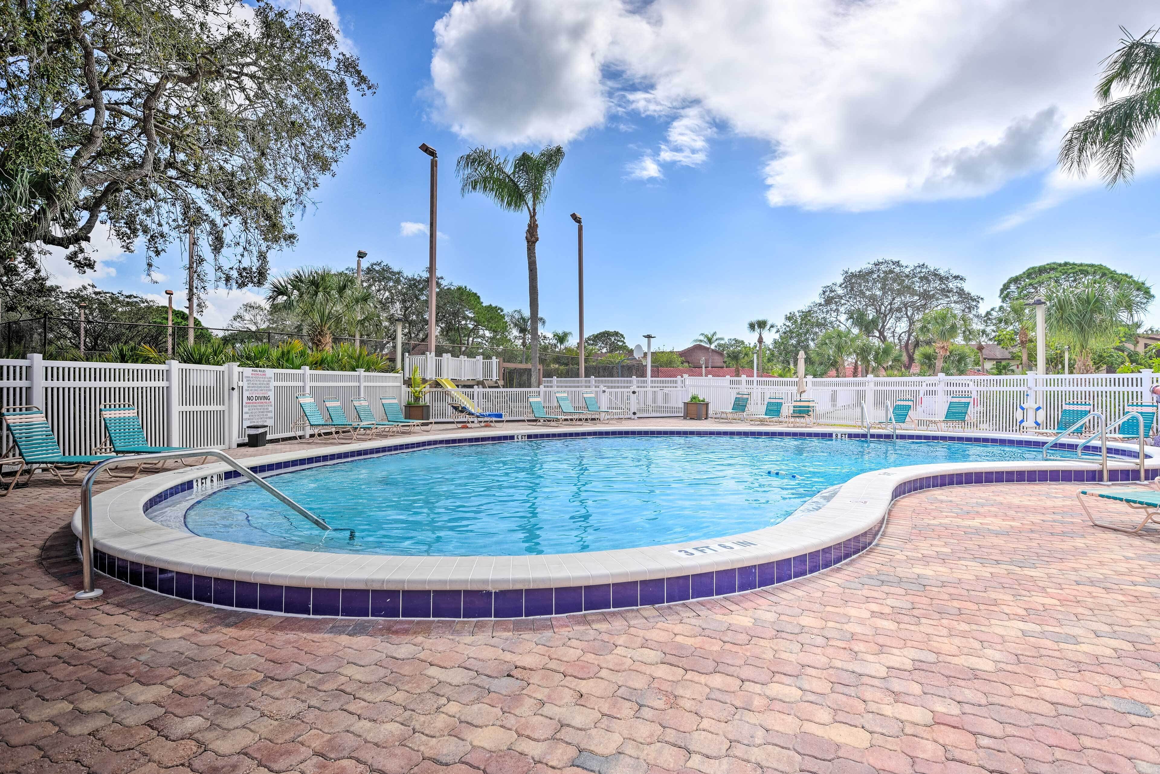 A swimming pool with a tiled edge, surrounded by a white fence and green lounge chairs.