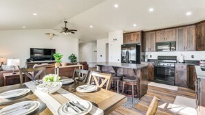 A modern kitchen with dark wooden cabinets, a central island, and a dining area with a wooden table set for four. - 2 Mi to Beaverhead River: Family-friendly Home! (Dillon)
