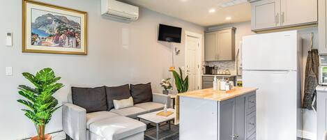 A modern living room with a sofa, coffee table, and a small kitchen area with a refrigerator and cabinets.