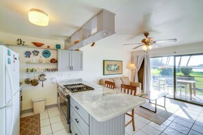 A kitchen with a central island, a dining area with a table and chairs, and a view of the outdoors through a sliding glass door. - Pool Access: Ocean-view Molokai Shores Condo! (Kaunakakai)