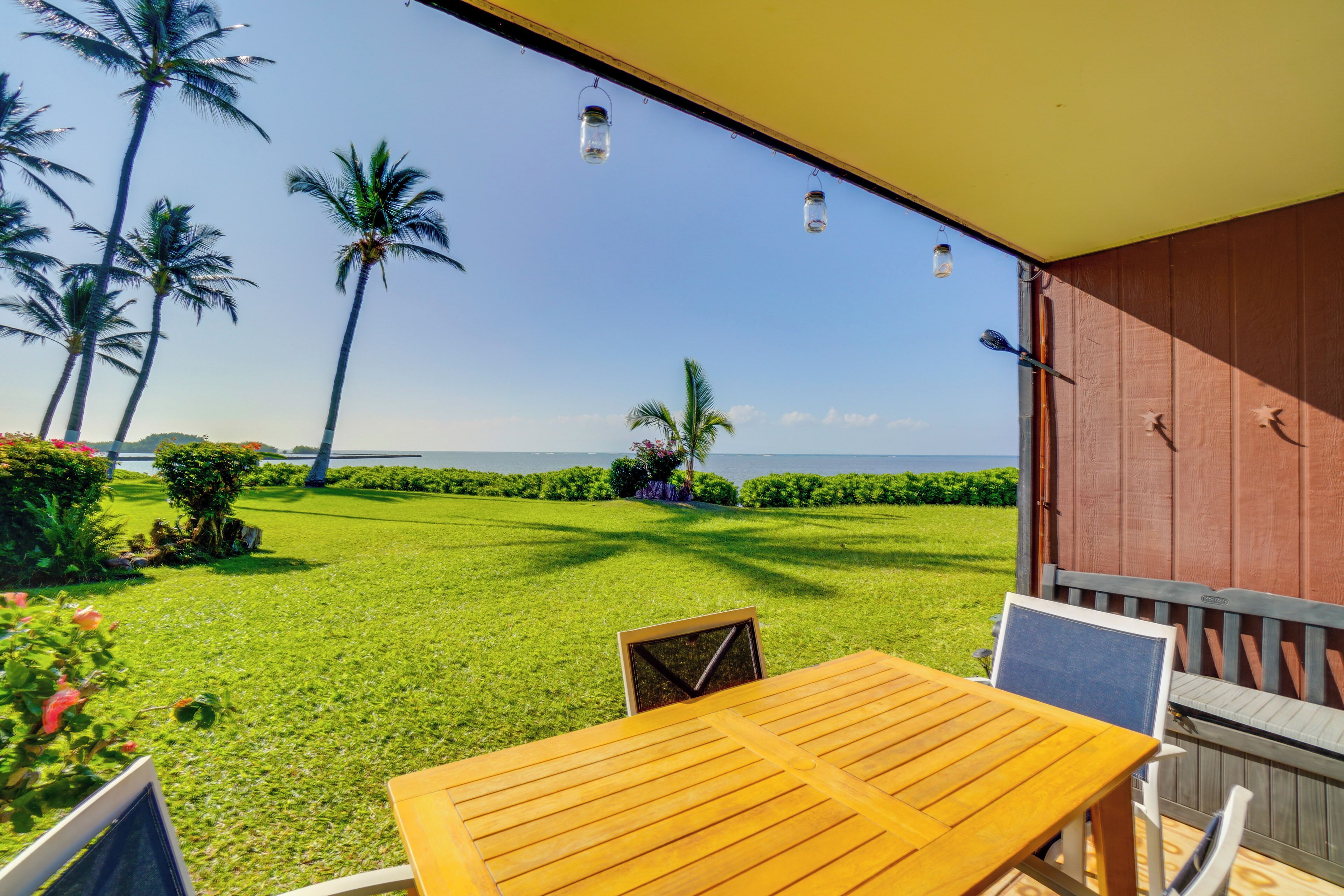 A wooden table and chairs on a balcony with a view of a grassy area, palm trees, and a distant body of water.