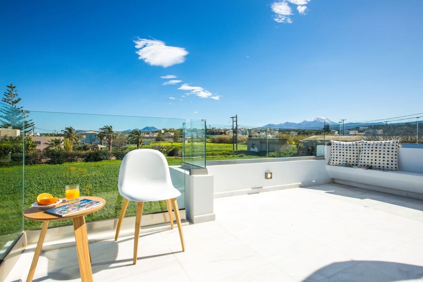 Eine Dachterrasse mit Glastrennwand, einem weißen Stuhl, einem kleinen Tisch mit einer Zeitschrift und Orangen sowie einer Couch mit gemusterten Kissen.