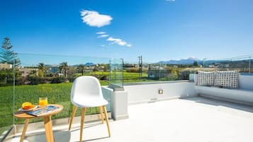 Eine Dachterrasse mit Glastrennwand, einem weißen Stuhl, einem kleinen Tisch mit einer Zeitschrift und Orangen sowie einer Couch mit gemusterten Kissen.
