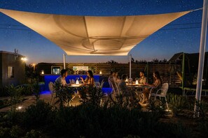 Un espace extérieur aménagé pour les repas, avec une table et des chaises sous une pergola, entouré de plantes et d’une piscine.