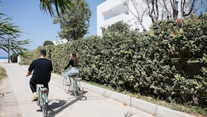 Dos personas andan en bicicleta por un camino pavimentado, junto a un seto y un edificio blanco.