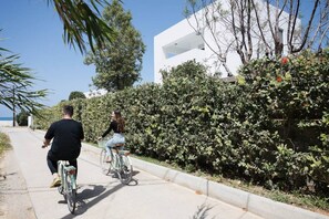 Two people riding bicycles on a paved path alongside a hedge and a white building. - Villa Melia bythe beach Thalasses villas by Domisi (Rethymno)