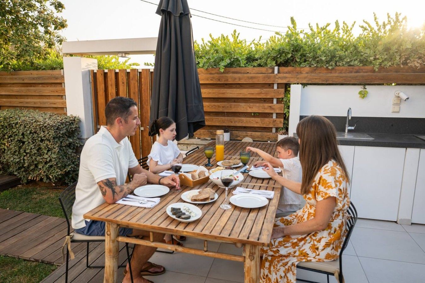 A family dining outdoors at a wooden table with a garden view, a barbecue grill, and a kitchen area.