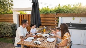 Una familia cenando al aire libre en una mesa de madera con vistas a un jardín, una parrilla y una zona de cocina.