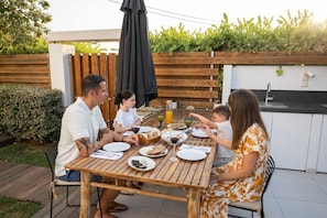 A family dining outdoors at a wooden table with a garden view, a barbecue grill, and a kitchen area. - Villa Melia bythe beach Thalasses villas by Domisi (Rethymno)