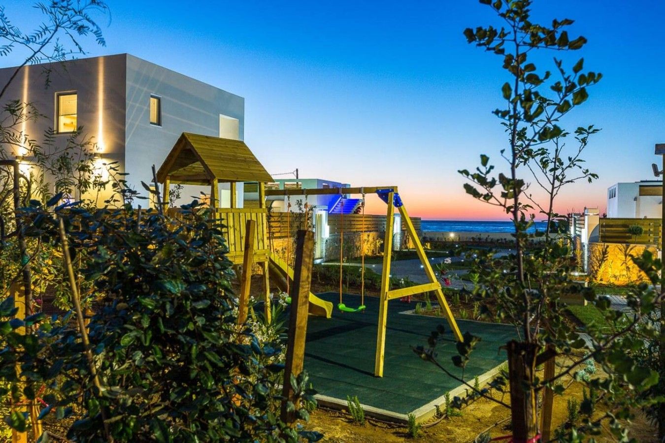 Ein modernes Wohngebiet mit einer Holzschaukel, einem Spielhäuschen und Blick auf das Meer im Abendlicht.