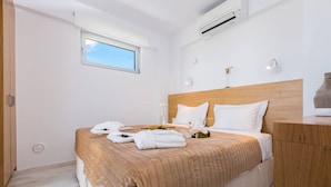 Habitación de hotel con cama, cabecera de madera, mesita de noche, ventana y equipo de aire acondicionado.