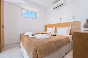 A hotel room with a bed, a wooden headboard, a nightstand, a window, and an air conditioning unit. - Villa Melia bythe beach Thalasses villas by Domisi (Rethymno)