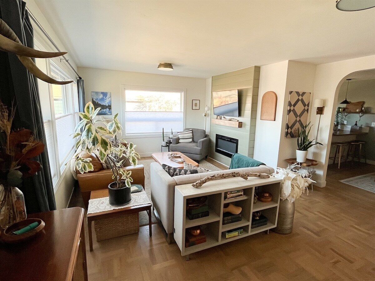 A living room with a sofa, coffee table, and a TV. There are plants, a fireplace, and a dining area visible.