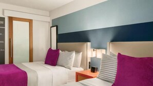 Une chambre d’hôtel avec deux lits, chacun recouvert d’une couverture et de coussins violets, une table de chevet avec une lampe et une porte en bois.