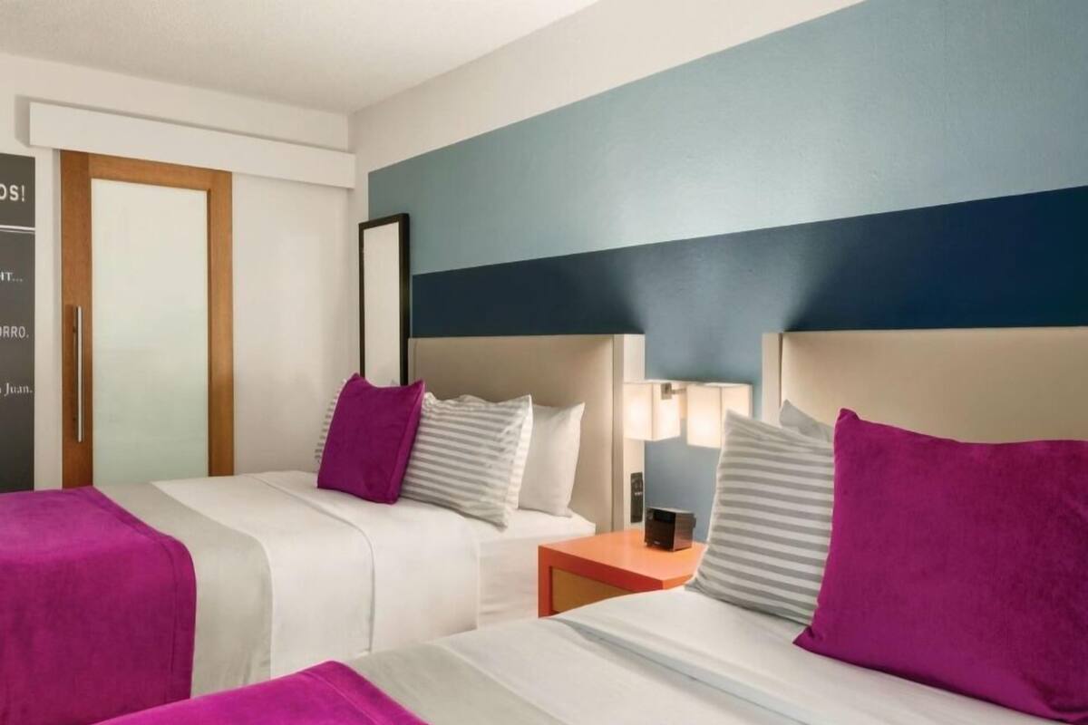 Habitación de hotel con dos camas, cada una con una manta y almohadas moradas, una mesita de noche con lámpara y una puerta de madera.