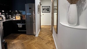Una cocina moderna con un refrigerador de acero inoxidable, suelo de madera en diseño de espiga y un jarrón blanco con flores secas en un estante.