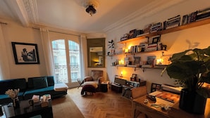 Una sala de estar con un sofá, un sillón y una mesita de centro. Se ven estanterías con libros y una planta.