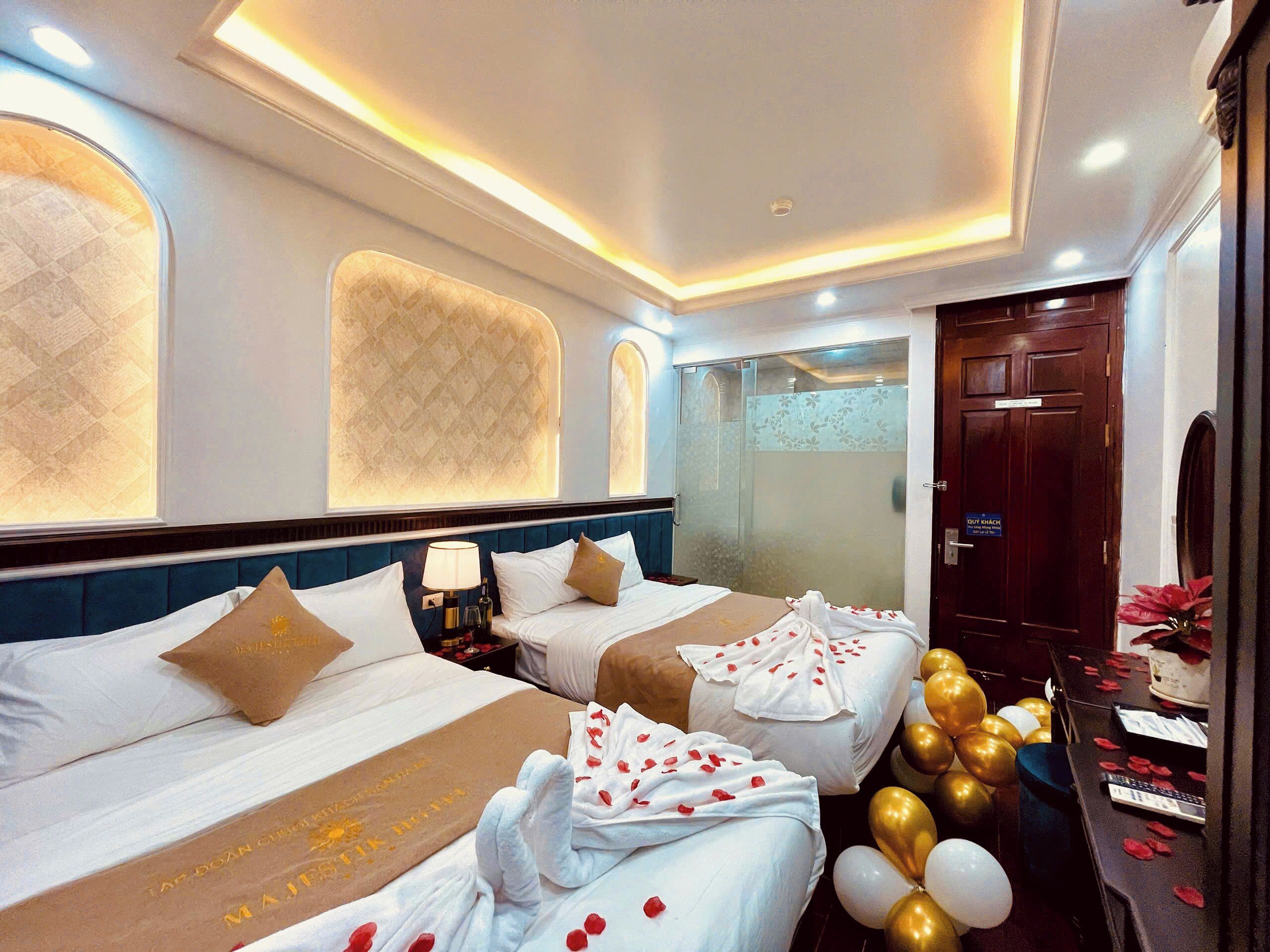 A hotel room with two beds, a minibar, a bathroom with a glass door, and a desk with balloons and rose petals.