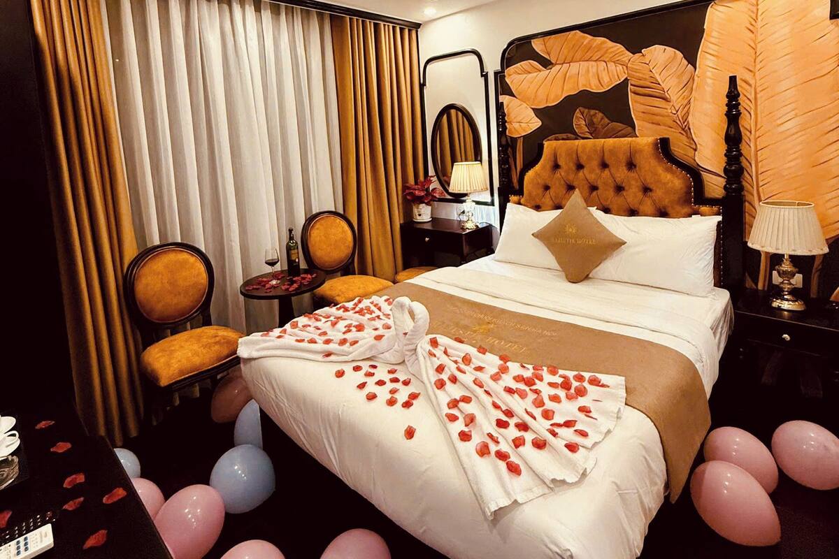 A hotel room with a large bed, a headboard with leaf designs, a nightstand, a chair, and a desk with a lamp.