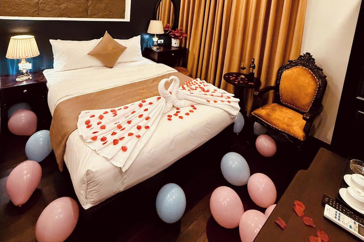 A hotel room with a large bed, a nightstand with a lamp, a chair, and balloons on the floor.