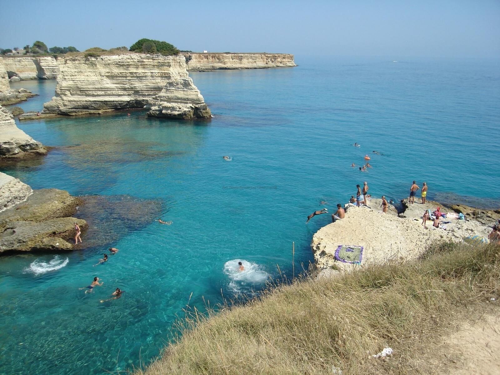A coastal area with clear blue water, rocky cliffs, and people swimming and sunbathing.