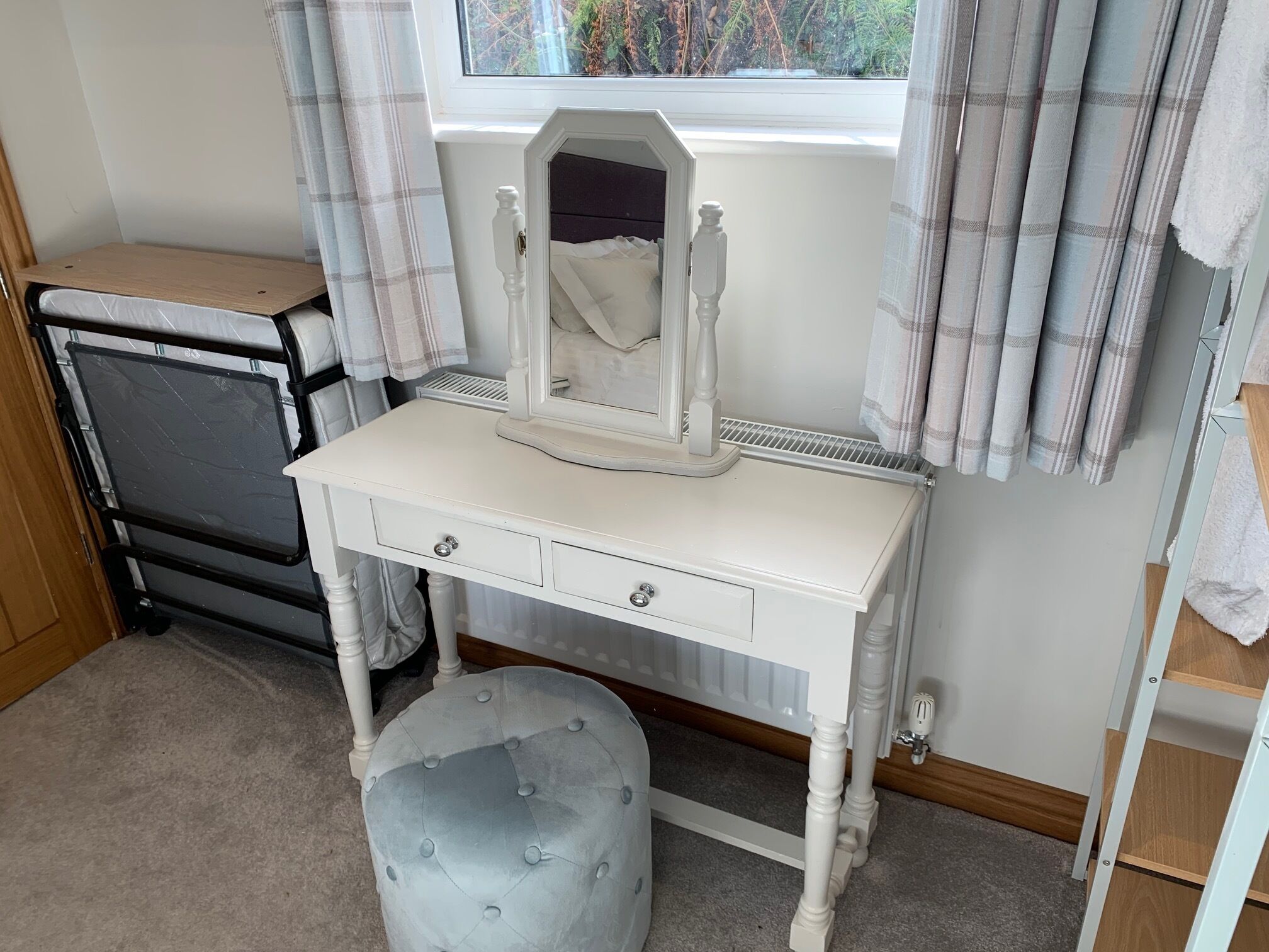 Une coiffeuse blanche avec un miroir, un petit tabouret et une fenêtre avec des rideaux.