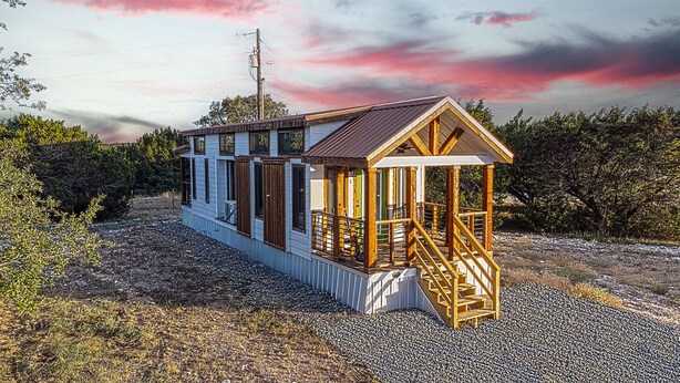 A small, single-story house with a porch and a staircase leading to a covered area. - Pet-Friendly Farmhouse Tiny Home w/ Private Deck (Lampasas)