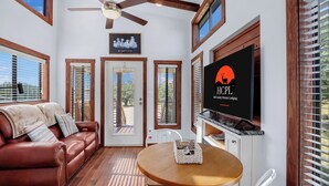 A cozy living room with a leather sofa, a wooden dining table, and a television displaying a logo. - Pet-Friendly Farmhouse Tiny Home w/ Private Deck (Lampasas)