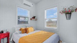 Une chambre avec un lit, deux fenêtres, une table de chevet rouge, un climatiseur fixé au mur et un pot de fleurs suspendu.