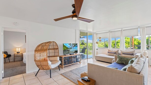 A spacious living room with a large window, a ceiling fan, a wicker chair, a sofa, and a TV stand.