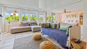 A spacious living room with a large sectional sofa, a smaller sofa, and a blue couch. There are two large windows with curtains, a floor lamp, and a ceiling fan.